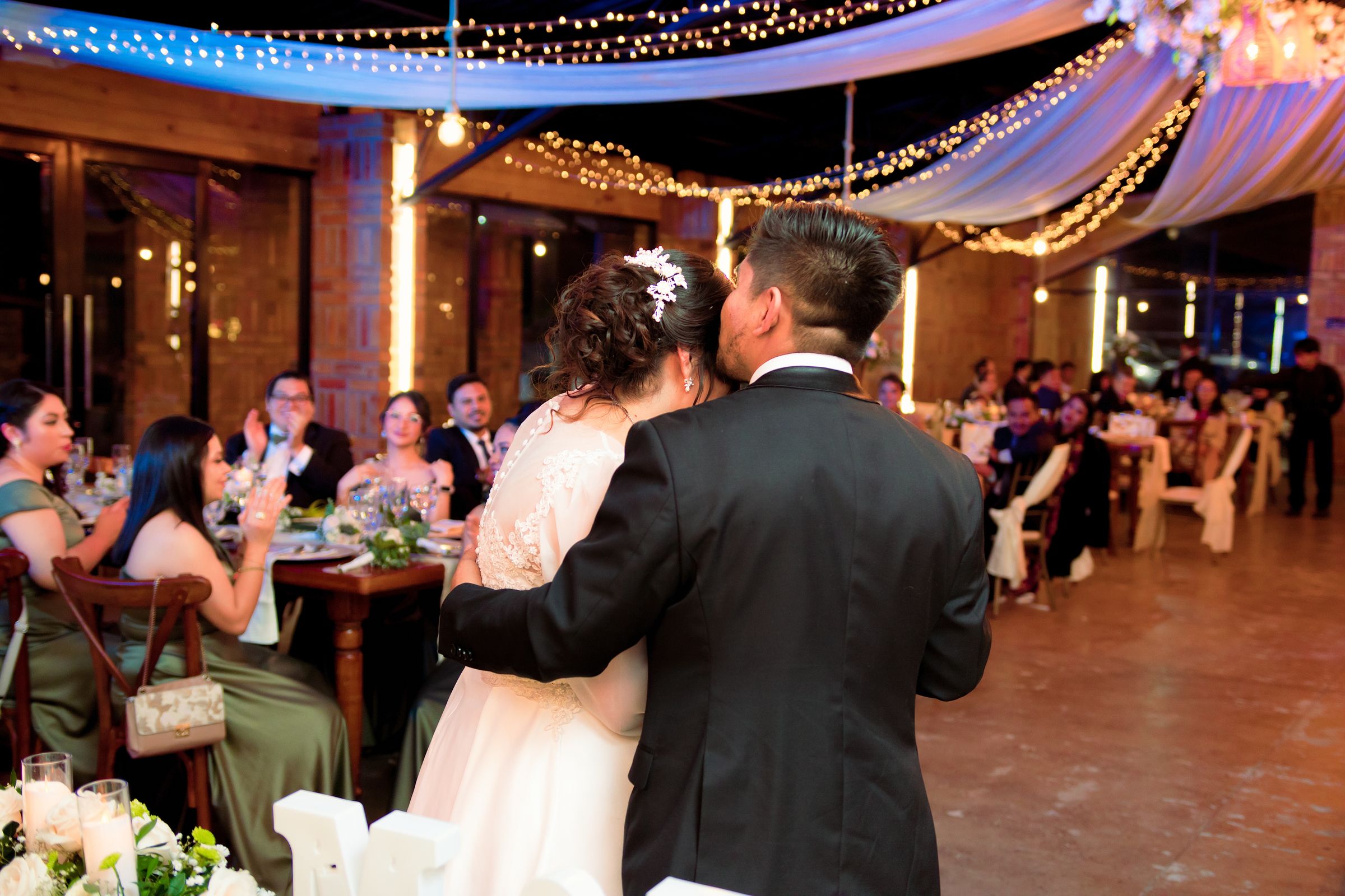 Novios abrazados en su recepción, con luces cálidas y los invitados aplaudiendo